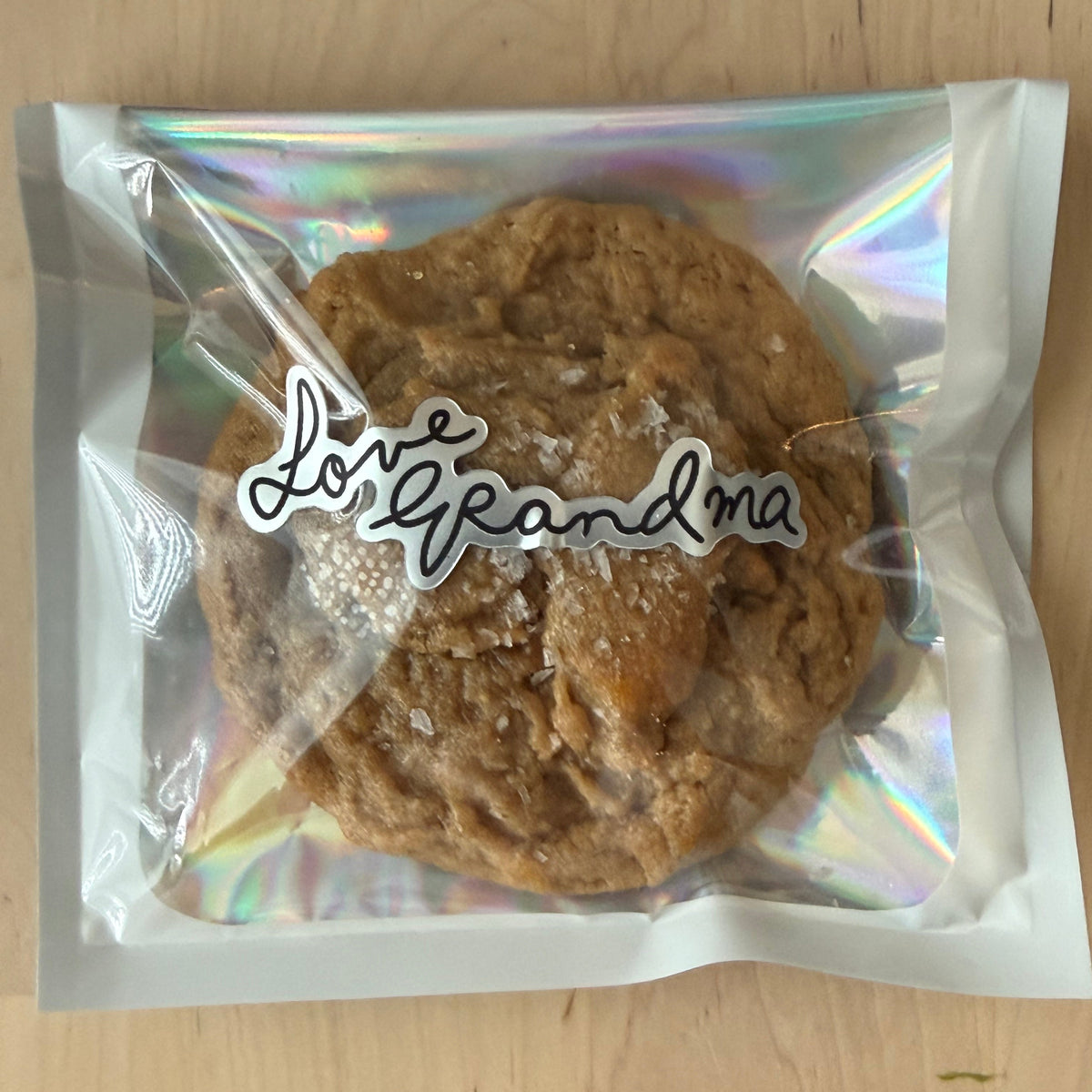A large chocolate chip cookie with coarse sea salt, sealed in a clear holographic bag with a white sticker reading 'Love Grandma' on a light wood tabletop.