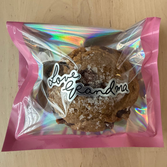 A baked cookie in a pink holographic zip bag with a white script logo reading 'day grandma', placed on a light wood tabletop.