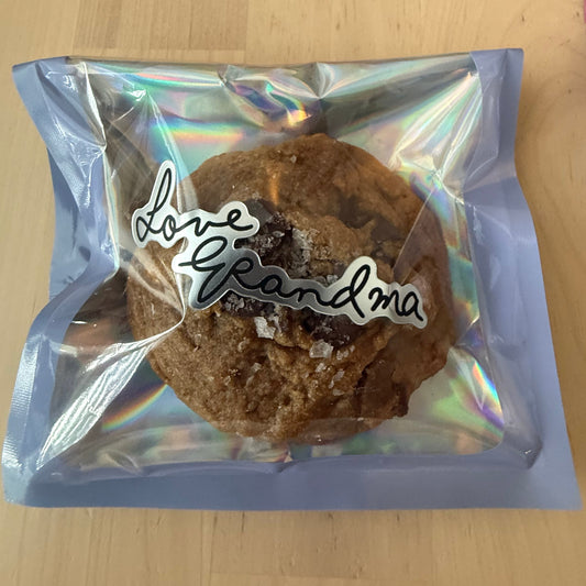 A chocolate chip cookie inside a clear holographic zip bag with the words 'love Grandma' printed on the front, placed on a light wood tabletop.