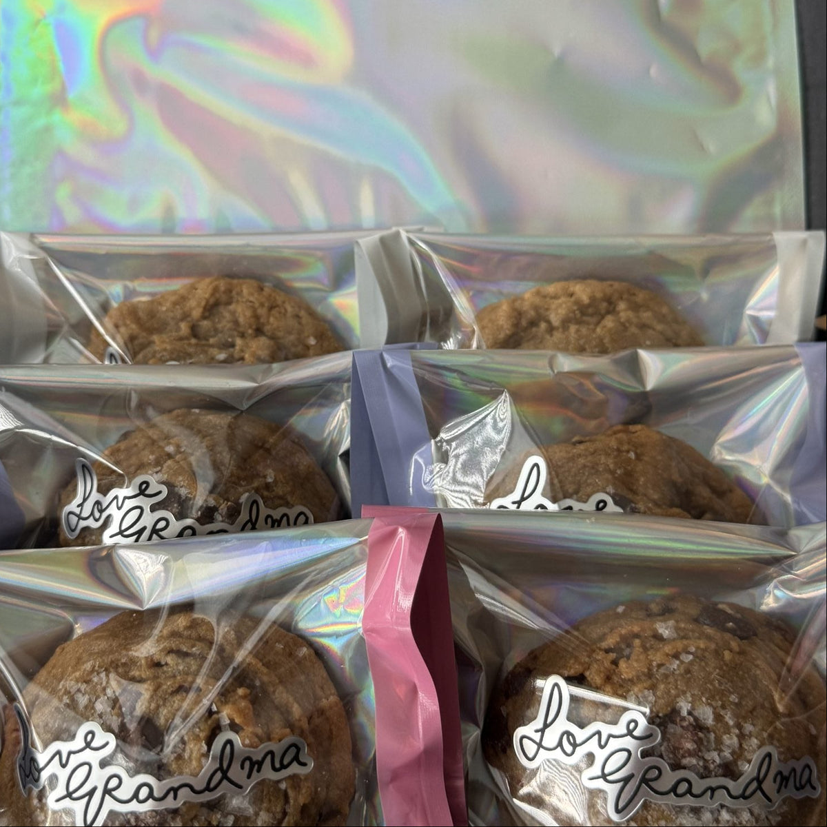 Several packaged cookies in clear bags with holographic silver seals and pink or lavender closures, arranged next to a matching iridescent pouch. The bags have a cursive script logo that appears to read 'J.