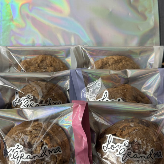 Several packaged cookies in clear bags with holographic silver seals and pink or lavender closures, arranged next to a matching iridescent pouch. The bags have a cursive script logo that appears to read 'J.