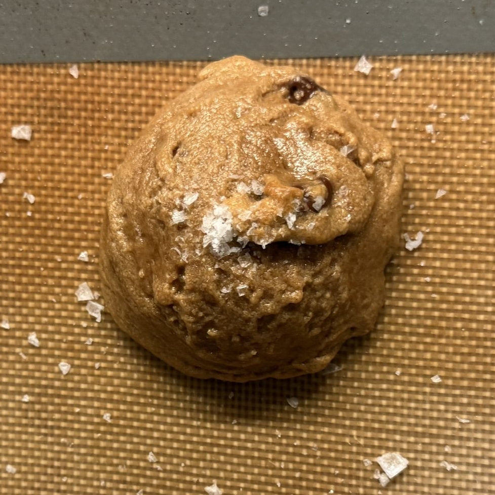 Unbaked chocolate chip cookies on a brown silicone baking mat, sprinkled with coarse Maldon sea salt