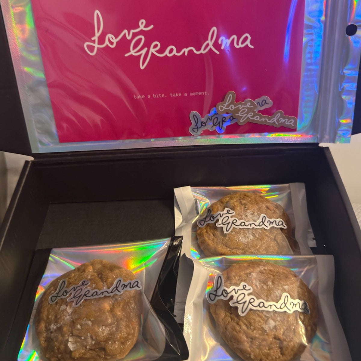An open black gift box containing three round brown cookies individually wrapped in holographic clear bags with 'Love Grandma' written on them, and a bright pink inner card with the same script.