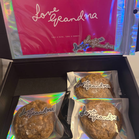 An open black gift box containing three round brown cookies individually wrapped in holographic clear bags with 'Love Grandma' written on them, and a bright pink inner card with the same script.